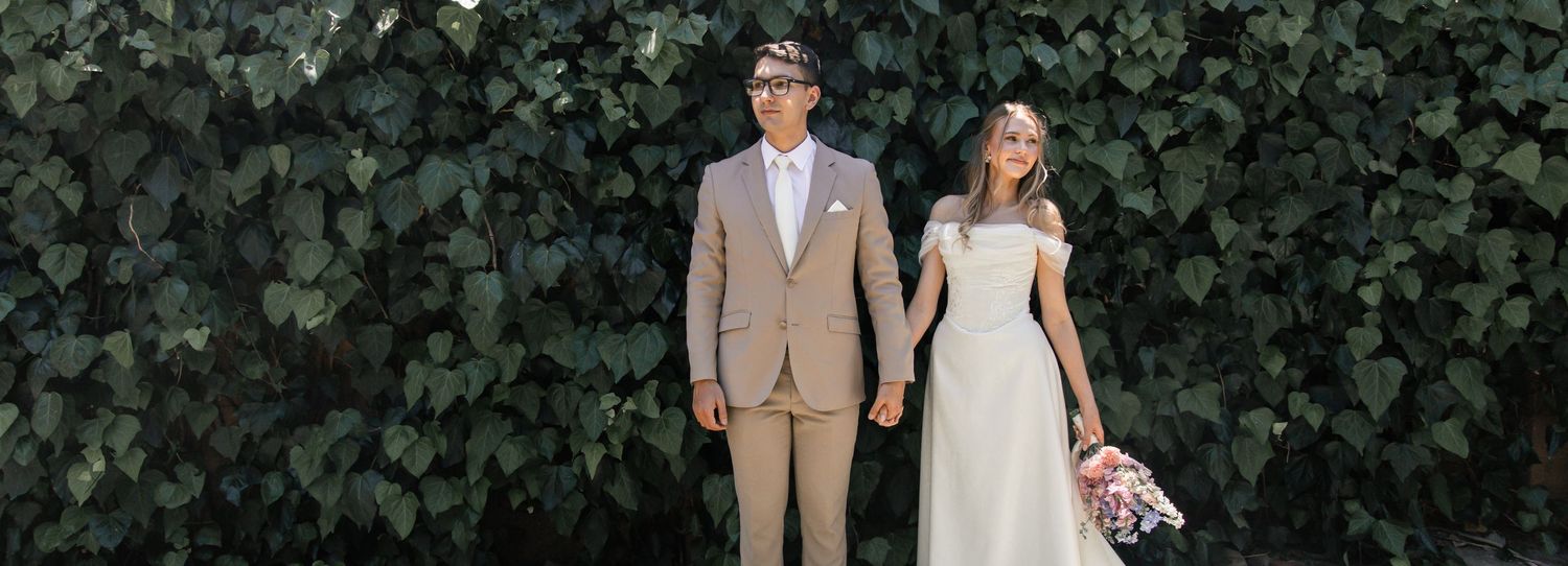 A couple in formal wedding attire poses for romantic photos in front of lush green foliage at an outdoor venue.