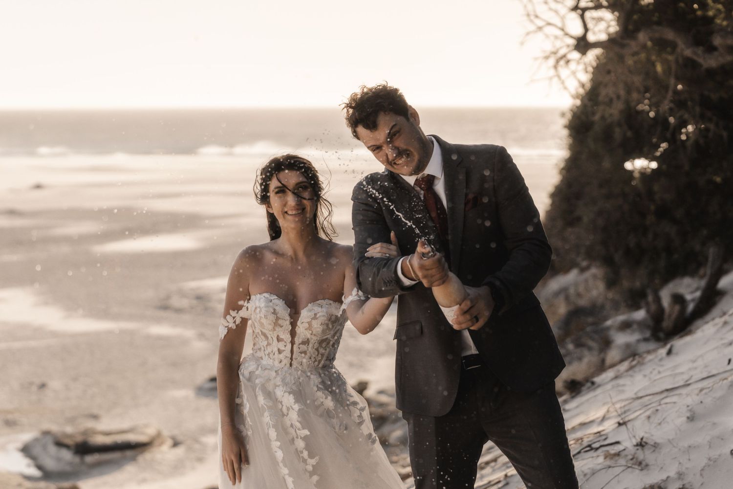 Bride and groom celebrate on a sandy beach, popping champagne in wedding attire with ocean waves in the background.