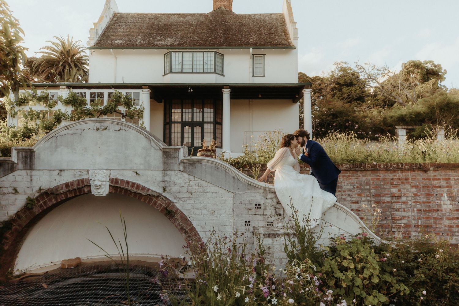 Bride and groom share a romantic kiss on a brick bridge in front of a charming white country house garden.