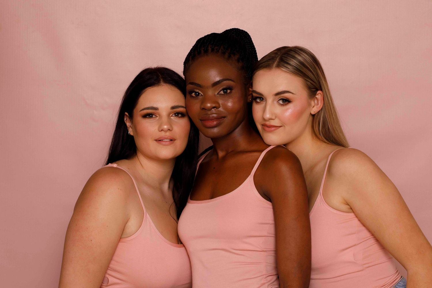 Diverse group of models wearing pink bras and underwear pose together against a pink background.