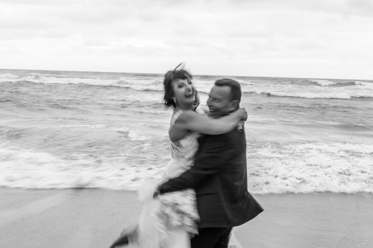 A couple dancing and embracing on a beach with waves crashing in the background, captured in black and white.