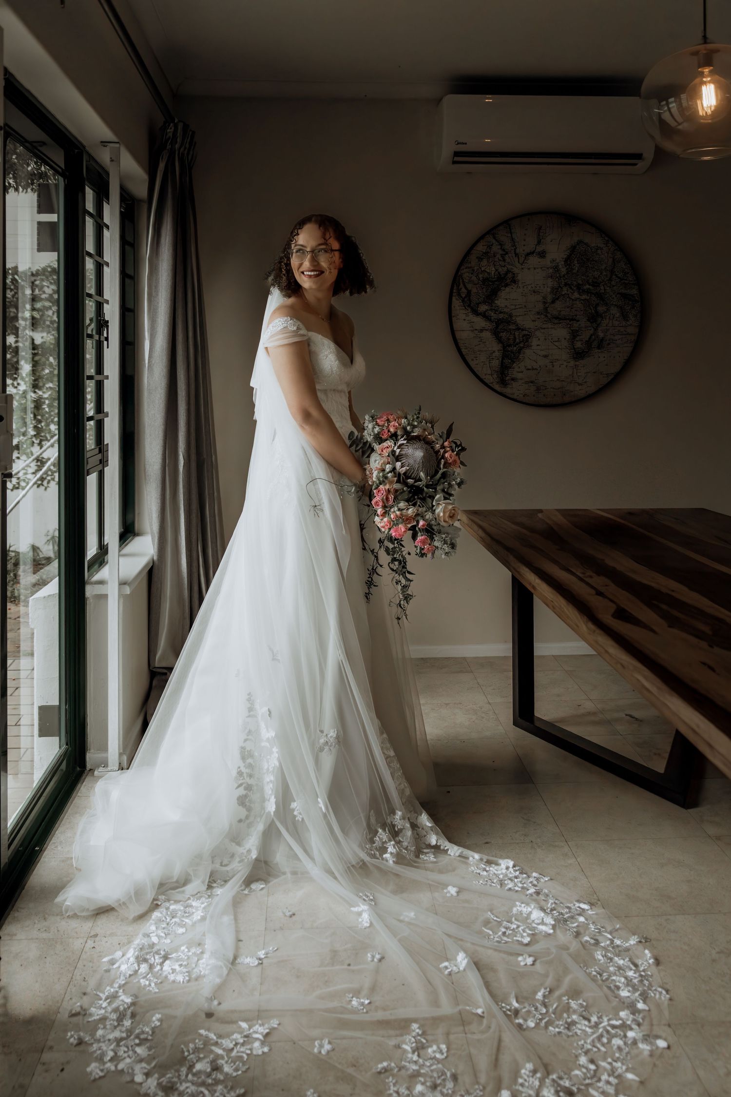 A bridal gown with intricate lace details and a flowing train is showcased in a modern indoor setting with natural light.