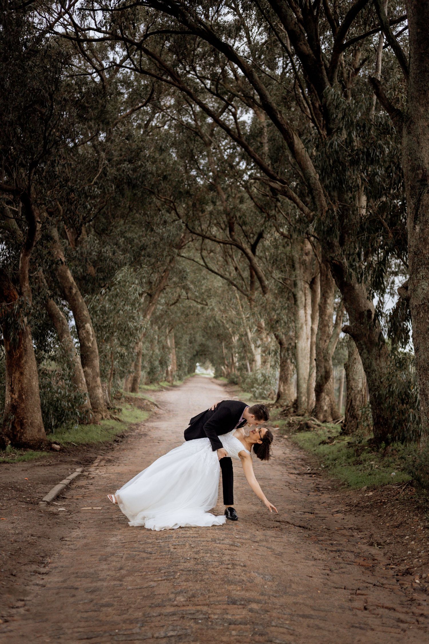 A romantic woodland path shows a couple performing dip pose in elegant wedding attire beneath a tunnel of trees.