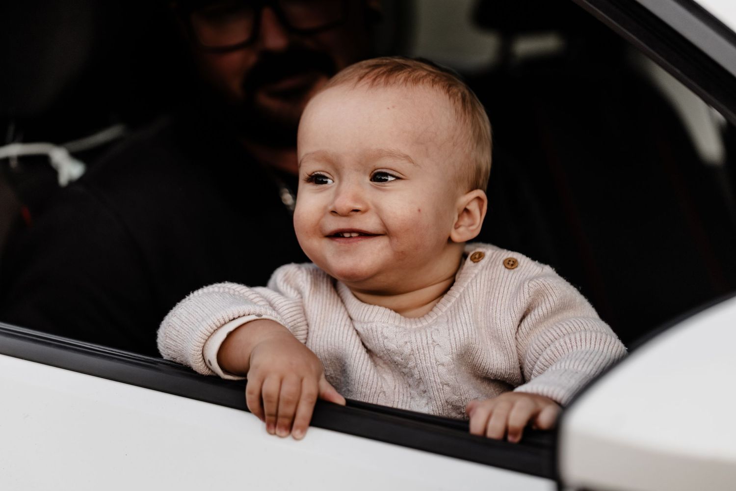 A happy baby wearing a light colored sweater peeks out from a car window with a cheerful smile.