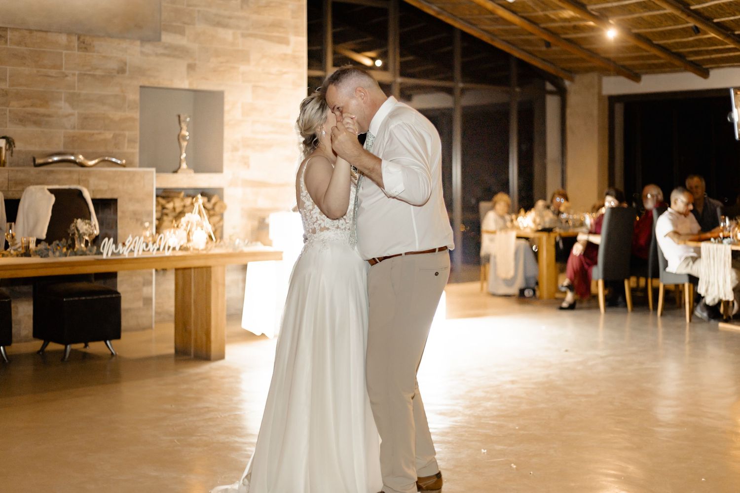 A romantic first dance between newlyweds at their wedding reception in a rustic venue with soft lighting and guests watching.
