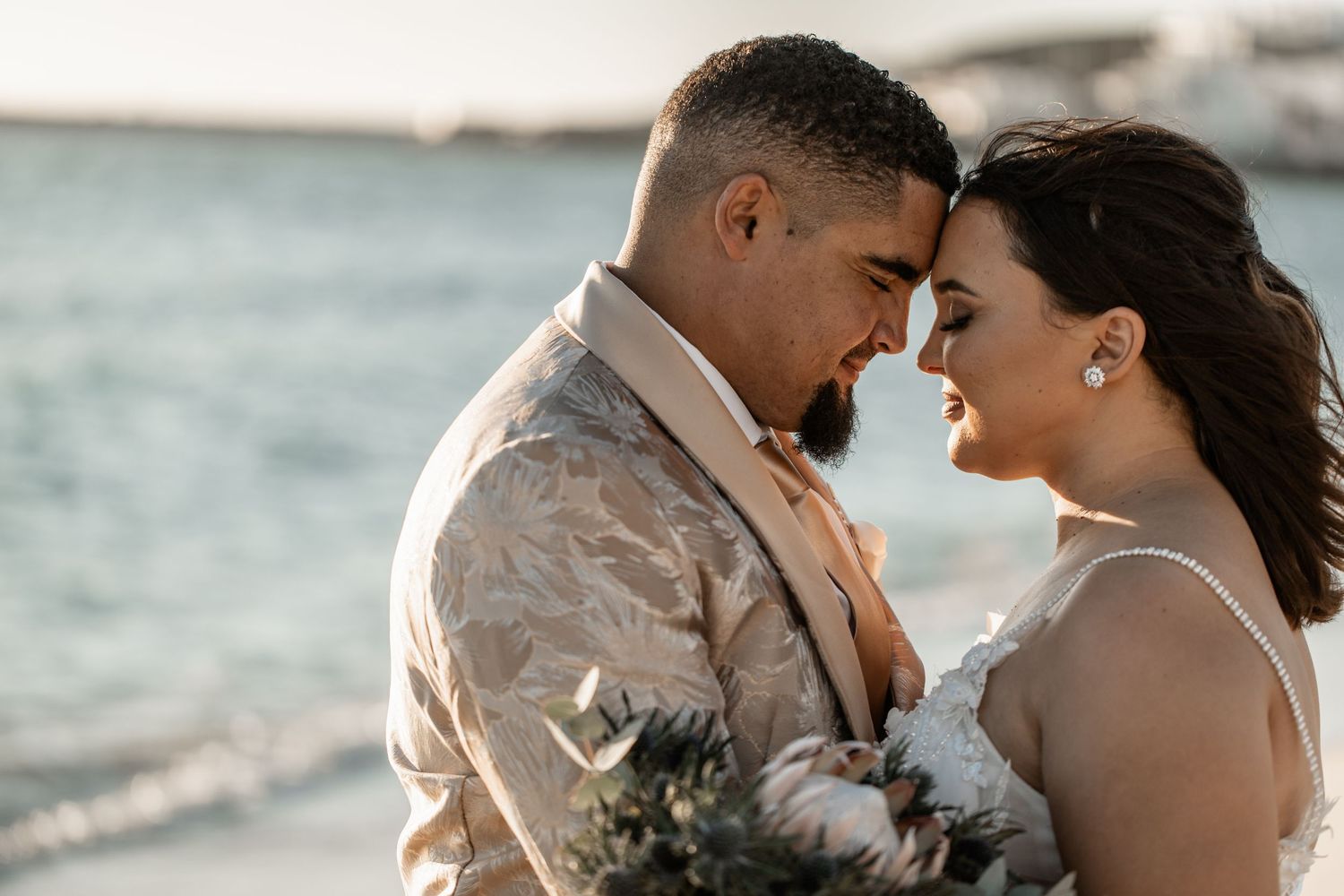A romantic newlywed couple shares an intimate moment at sunset by the ocean, with the bride holding a floral bouquet.