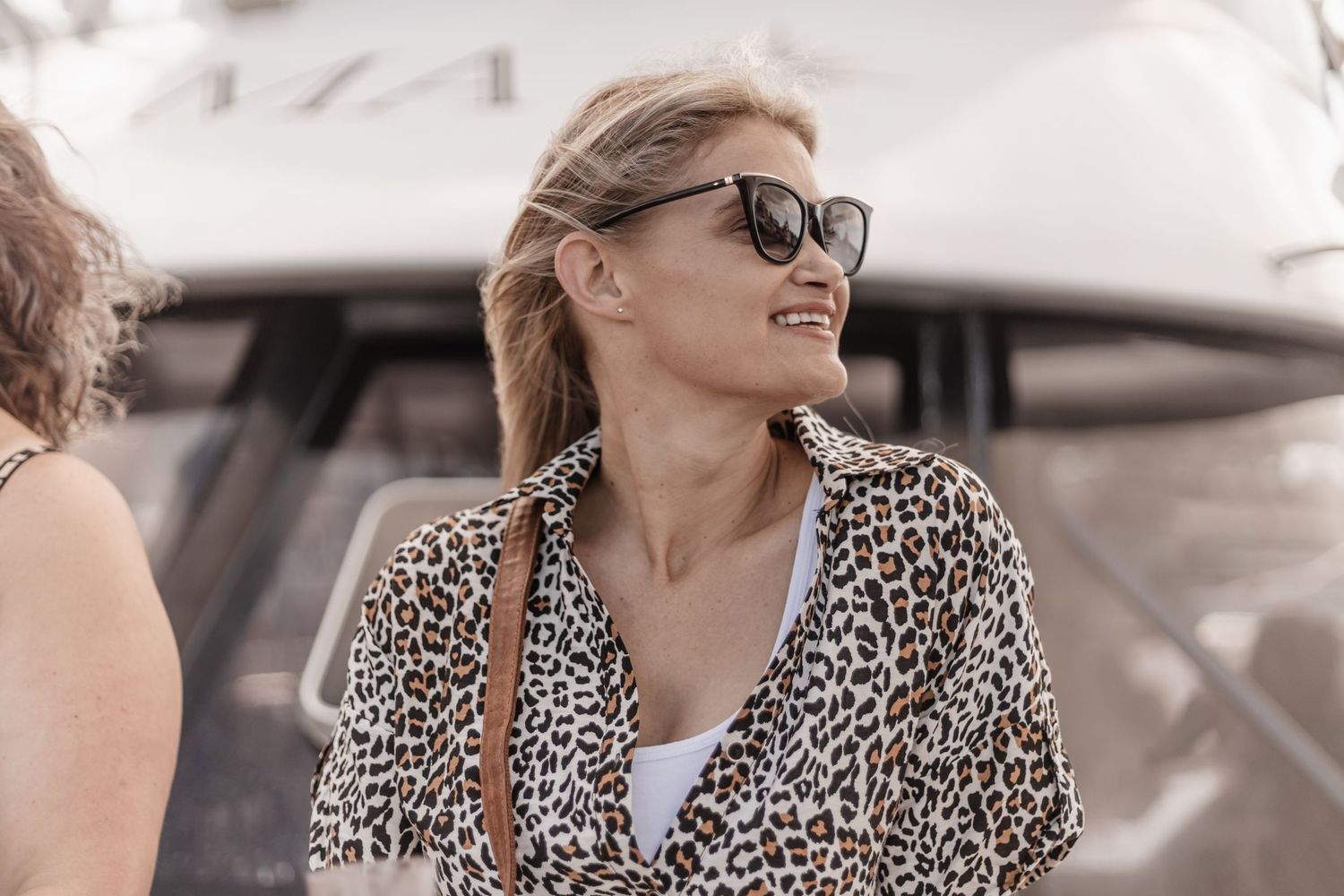 Fashionable person in leopard print jacket and sunglasses smiling while outside on sunny day.