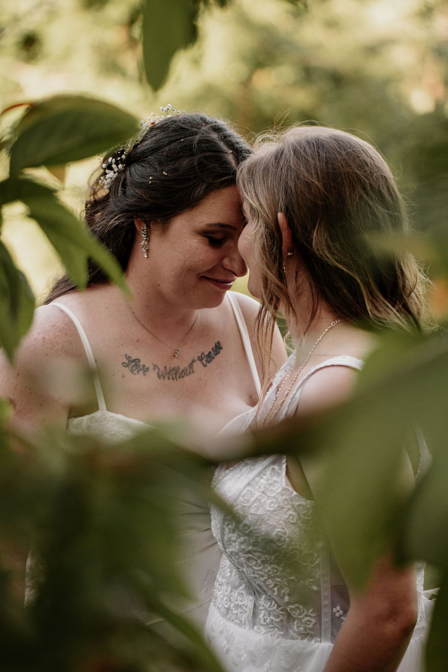 An intimate wedding moment captured through green foliage, showing two people in white dresses sharing a tender embrace.
