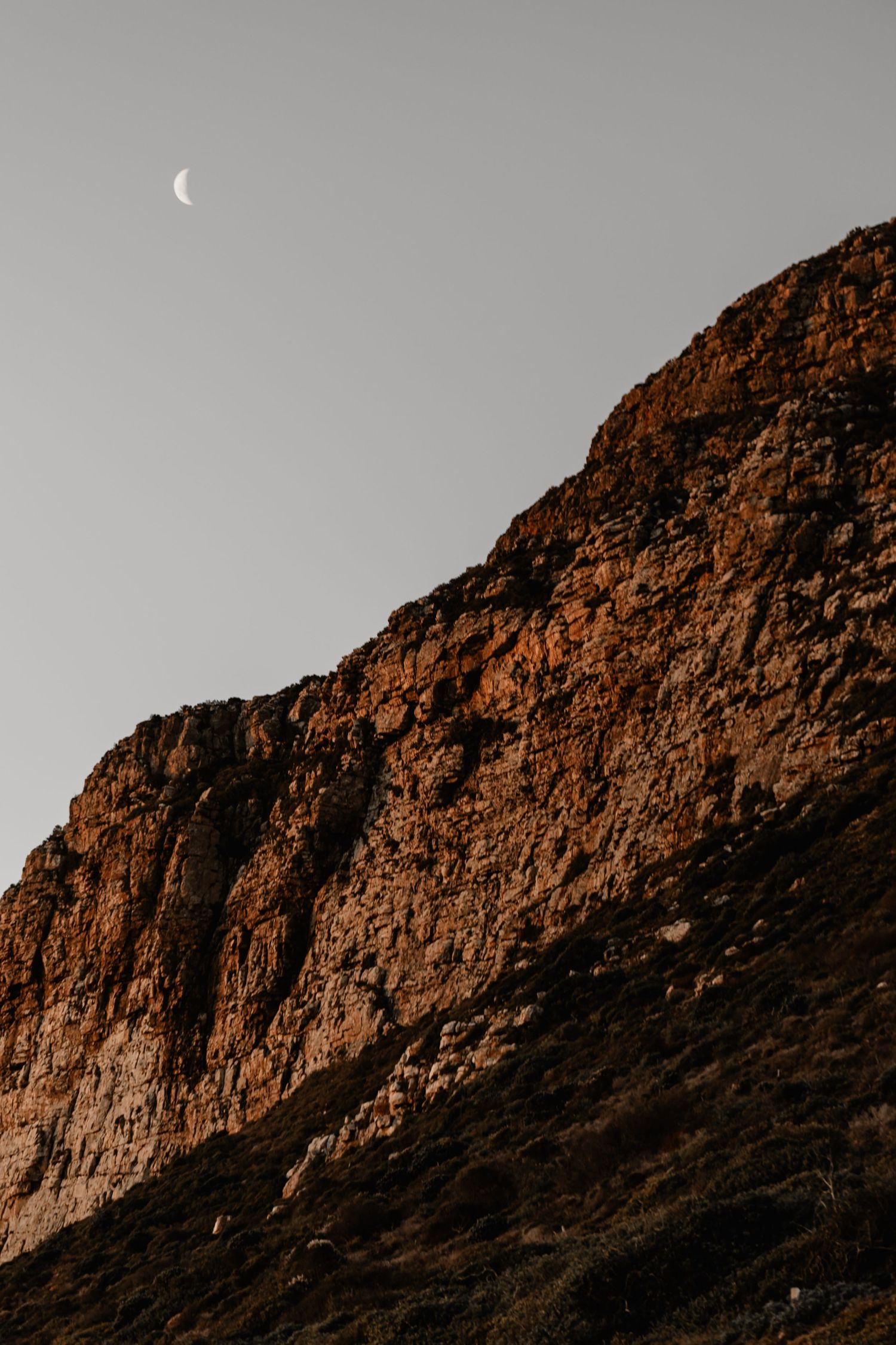 Dramatic brown rocky mountain cliff face with a crescent moon visible in the gray evening sky.