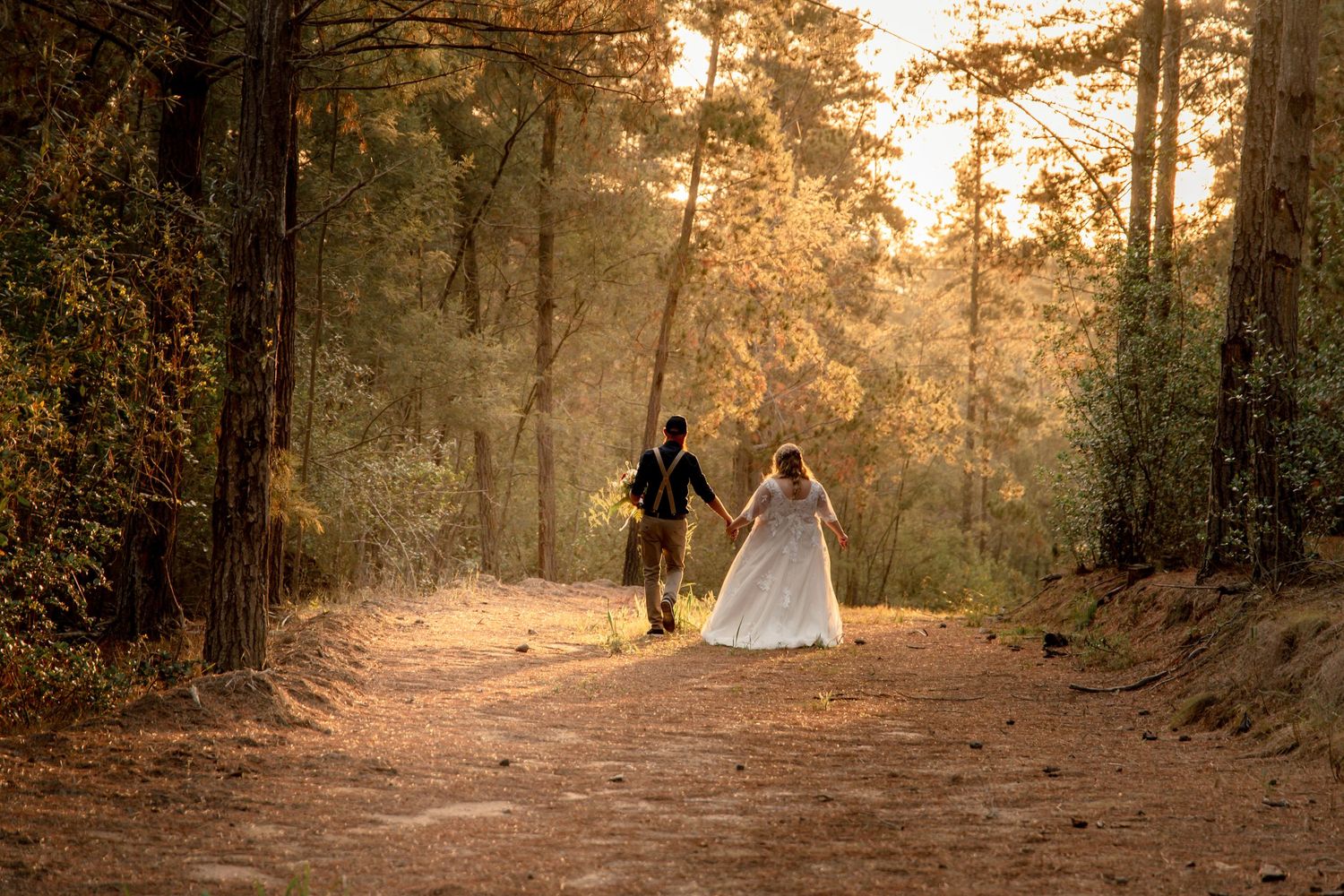 Bride and groom holding hands walking down a sunlit forest path in wedding attire.