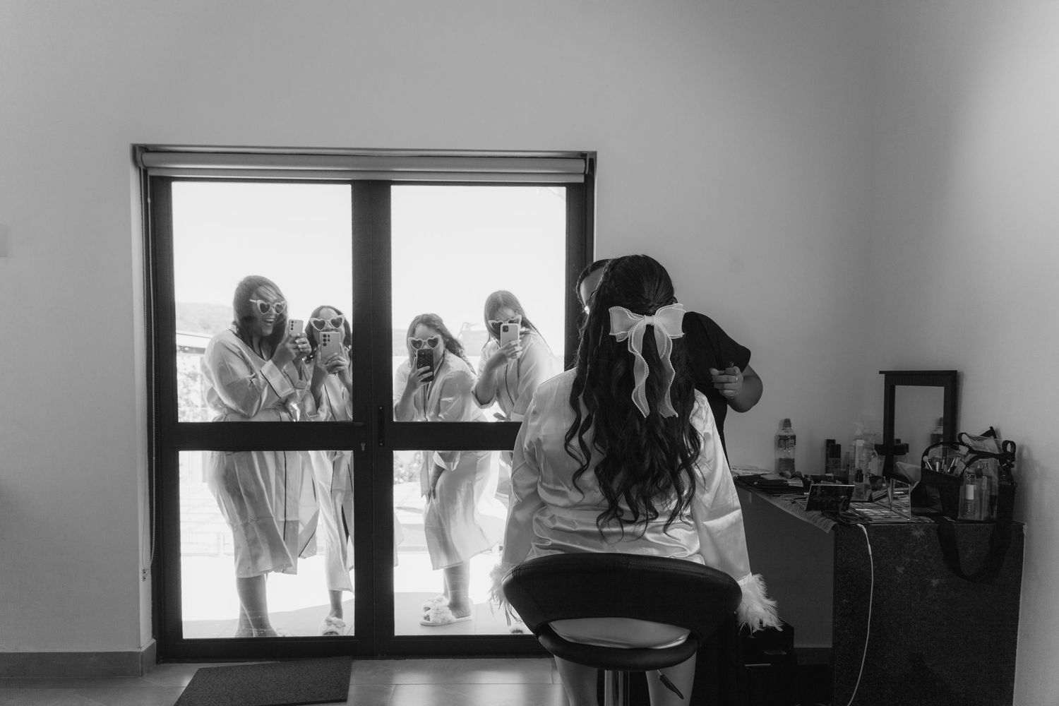 Bride sits at vanity while bridesmaids peek through glass door in surprise, captured in black and white wedding photo.