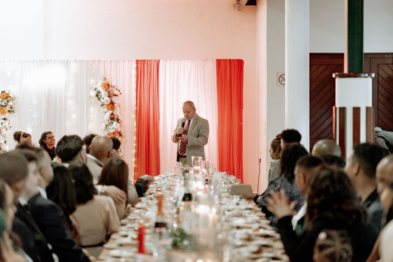 Event guests mingle in an elegant venue with coral curtains and white tablecloths during a formal gathering.