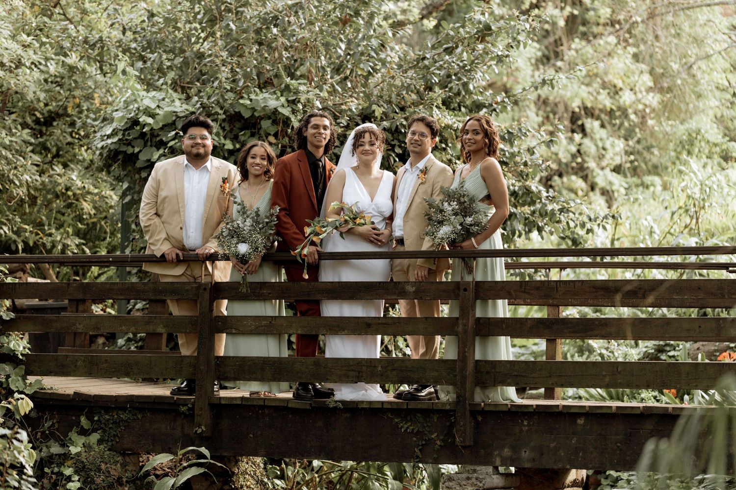Wedding party gathers along a rustic wooden bridge in a sunlit forest setting for outdoor photos.