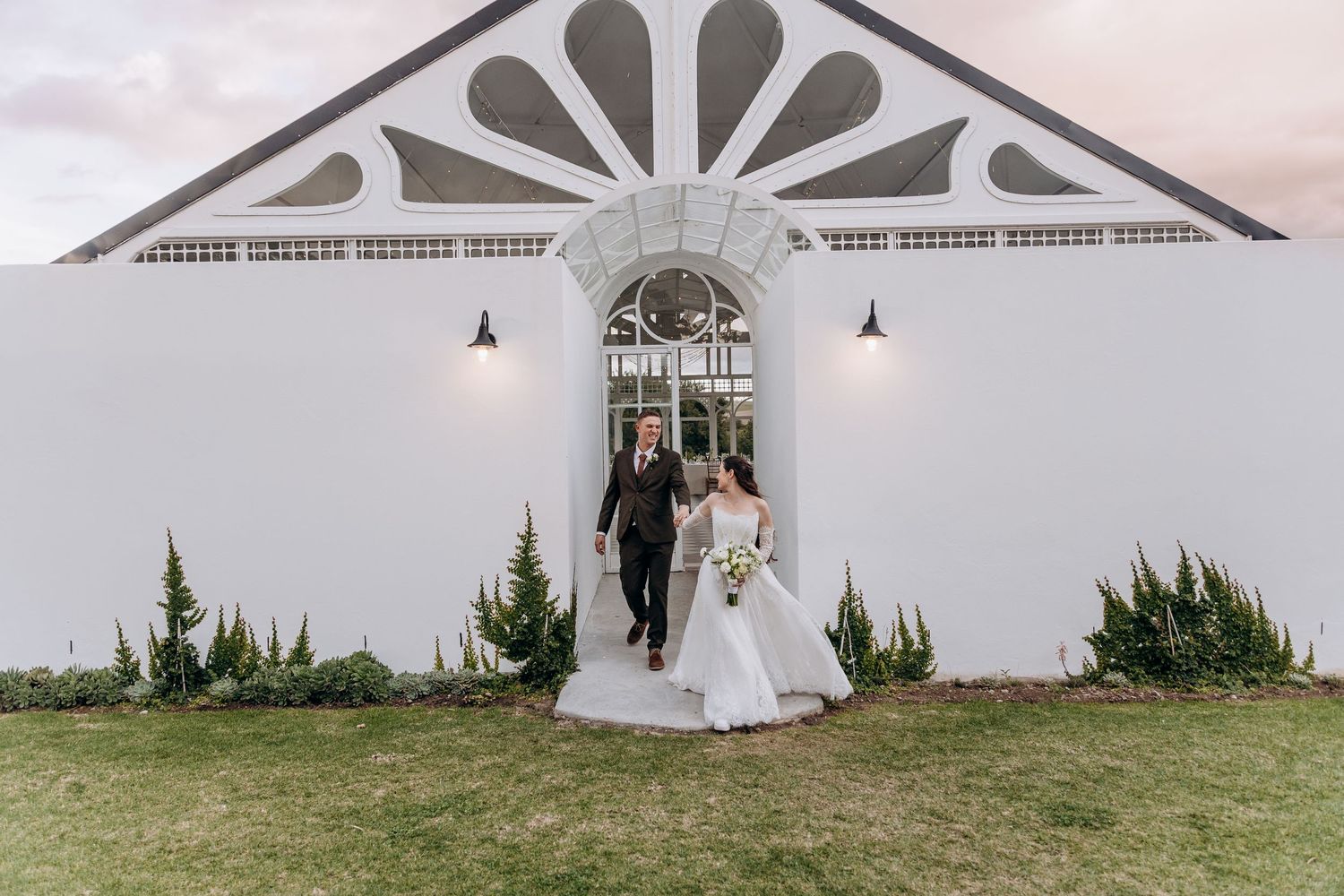 Bride and groom walking hand in hand outside a white chapel wedding venue at dusk.