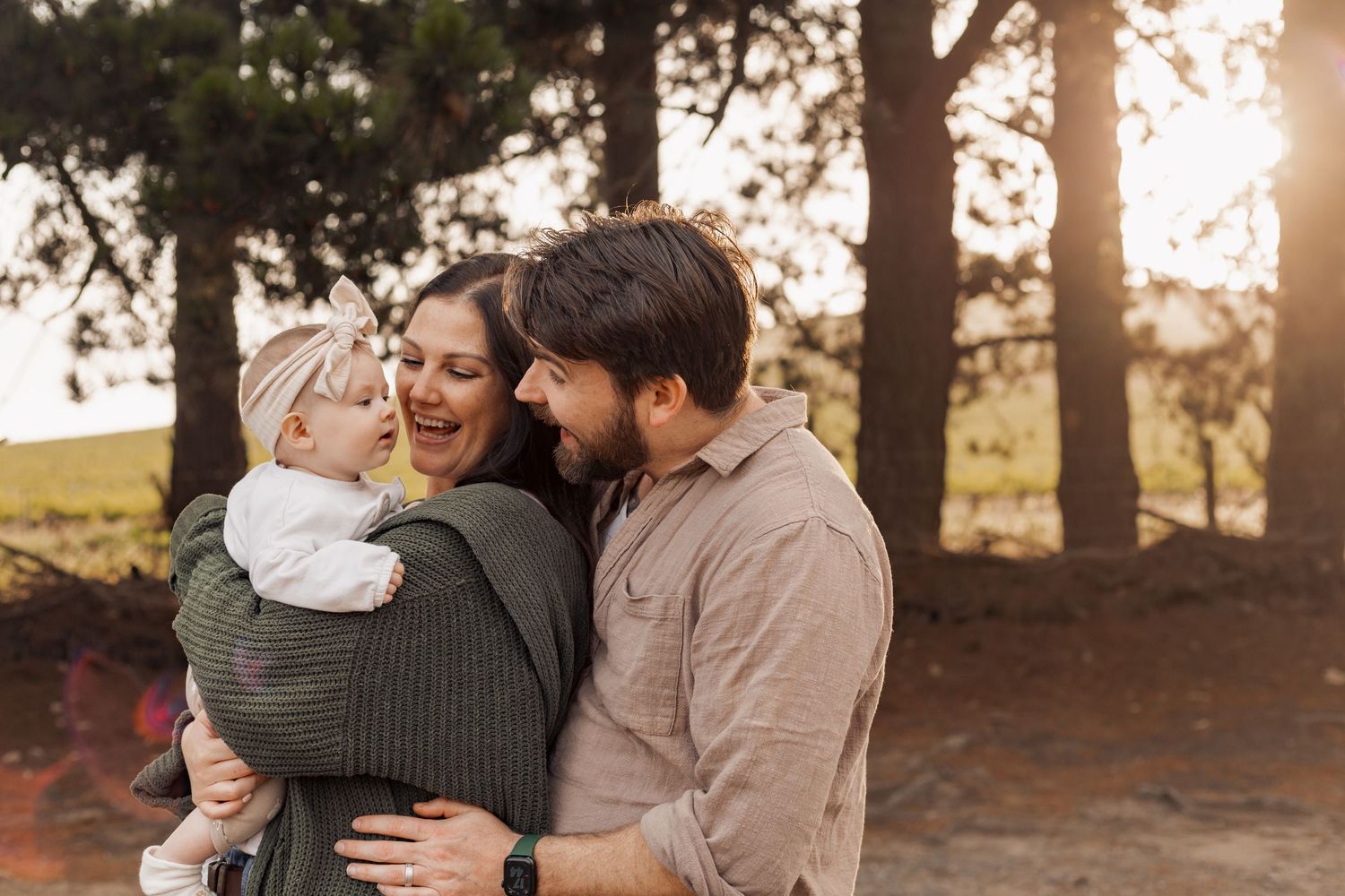 A loving family embraces outdoors during golden hour with tall pine trees in the background.