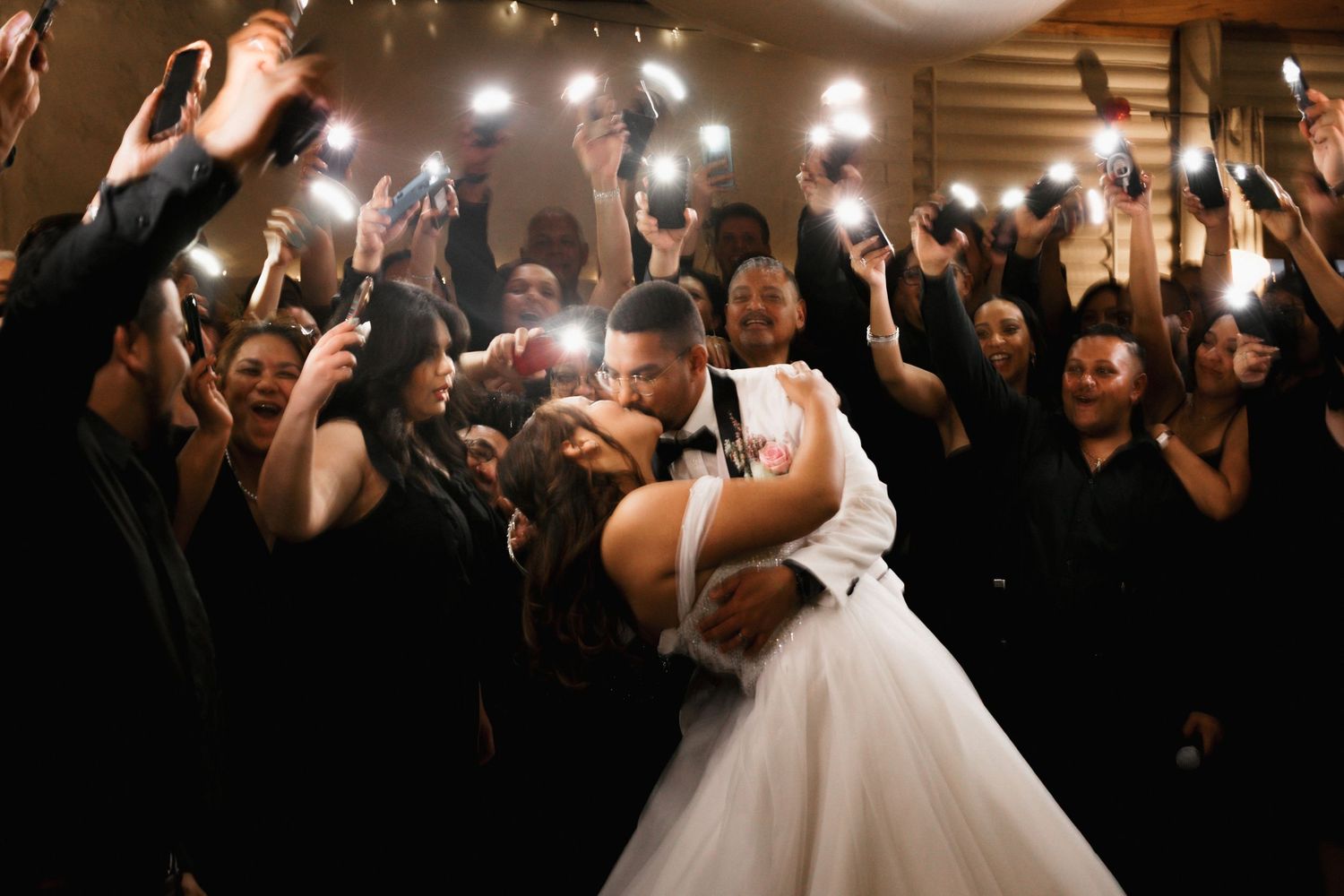 A couple shares their first dance at a wedding reception while guests watch and take photos with their phones.