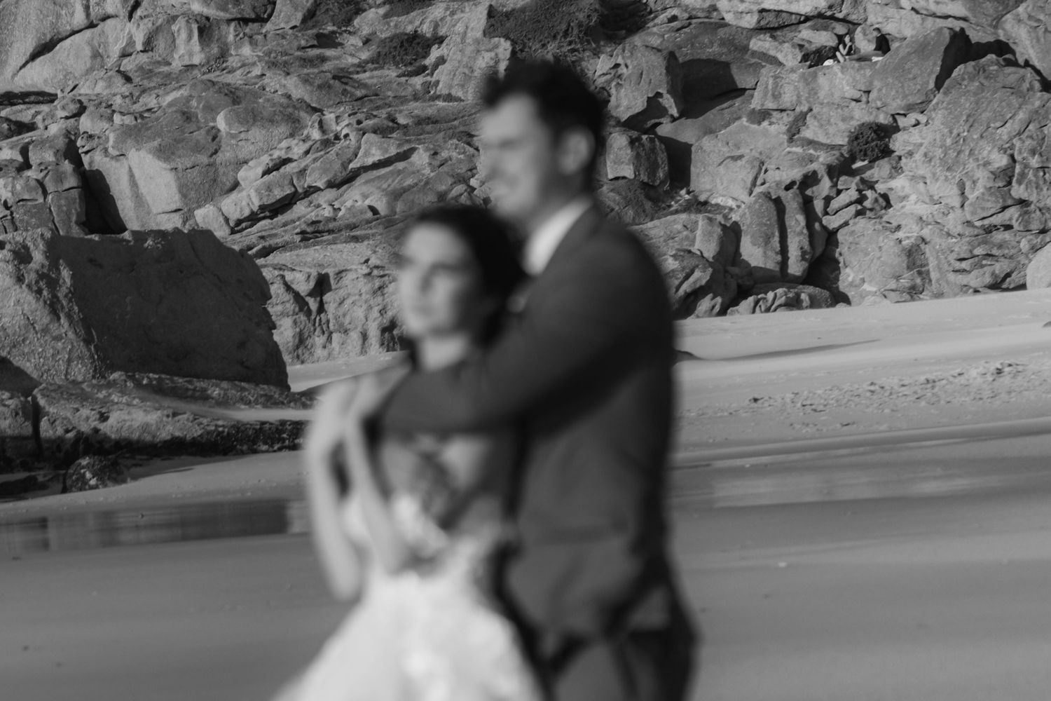 Black and white photo of a couple embracing on a beach with rocky cliffs, dressed in wedding attire.