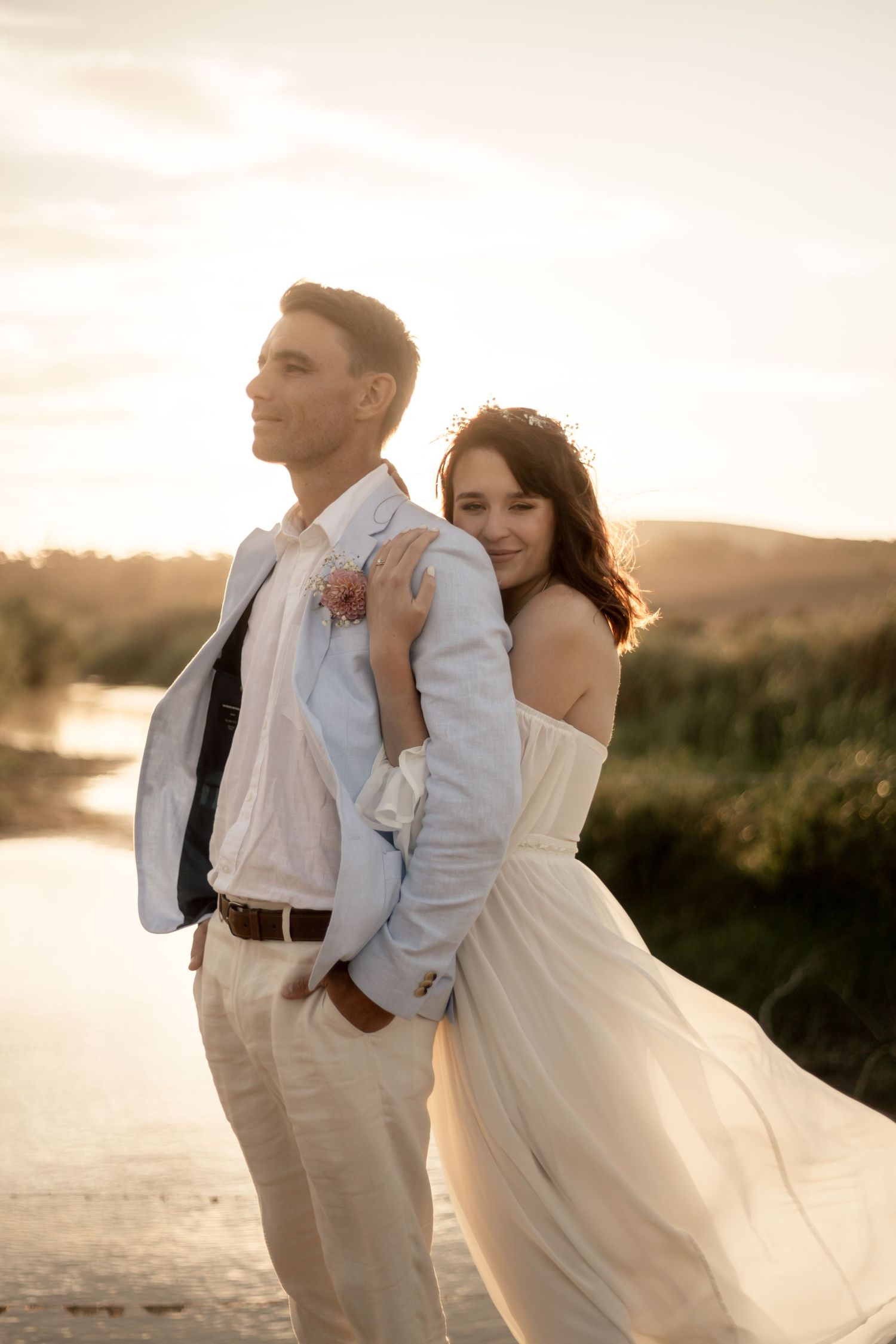 Couple in wedding attire embracing at sunset near a river, bride in white off-shoulder gown hugging groom in light suit.