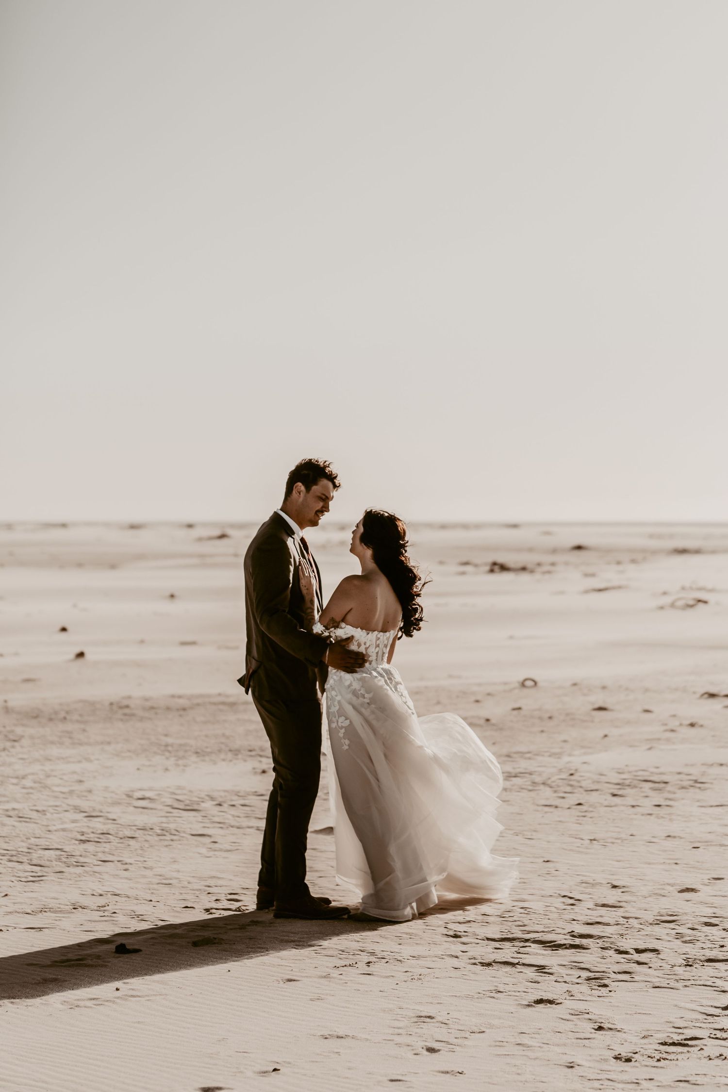 Bride and groom holding hands on a sandy beach, bride wearing a flowing white off-shoulder gown in a romantic outdoor setting.