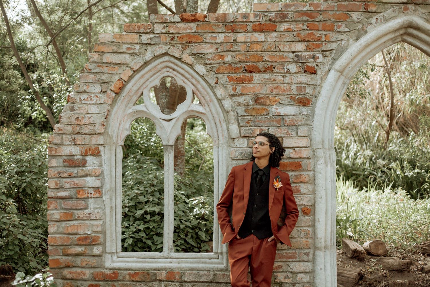 Someone in a brown coat poses near rustic brick walls and arched windows in a garden setting.