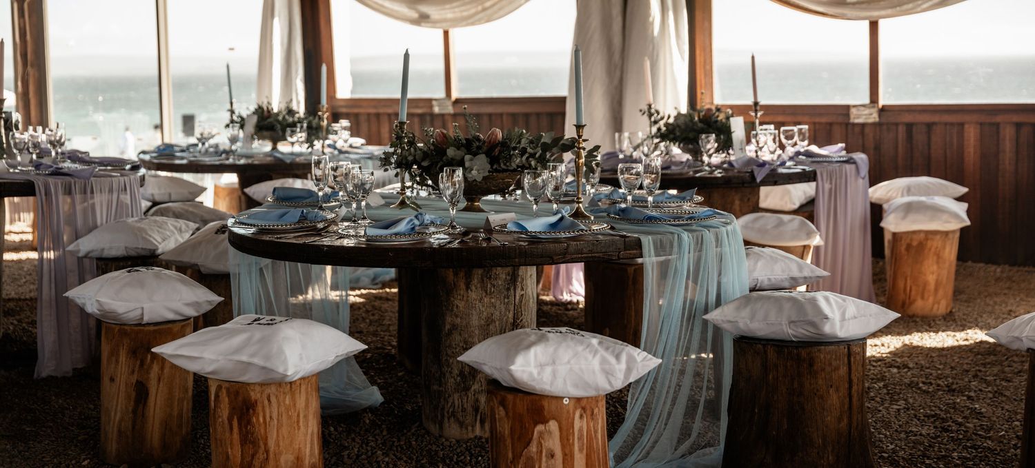 A rustic wedding reception table setup with tree stump seating and teal accents in a windowed venue overlooking water.