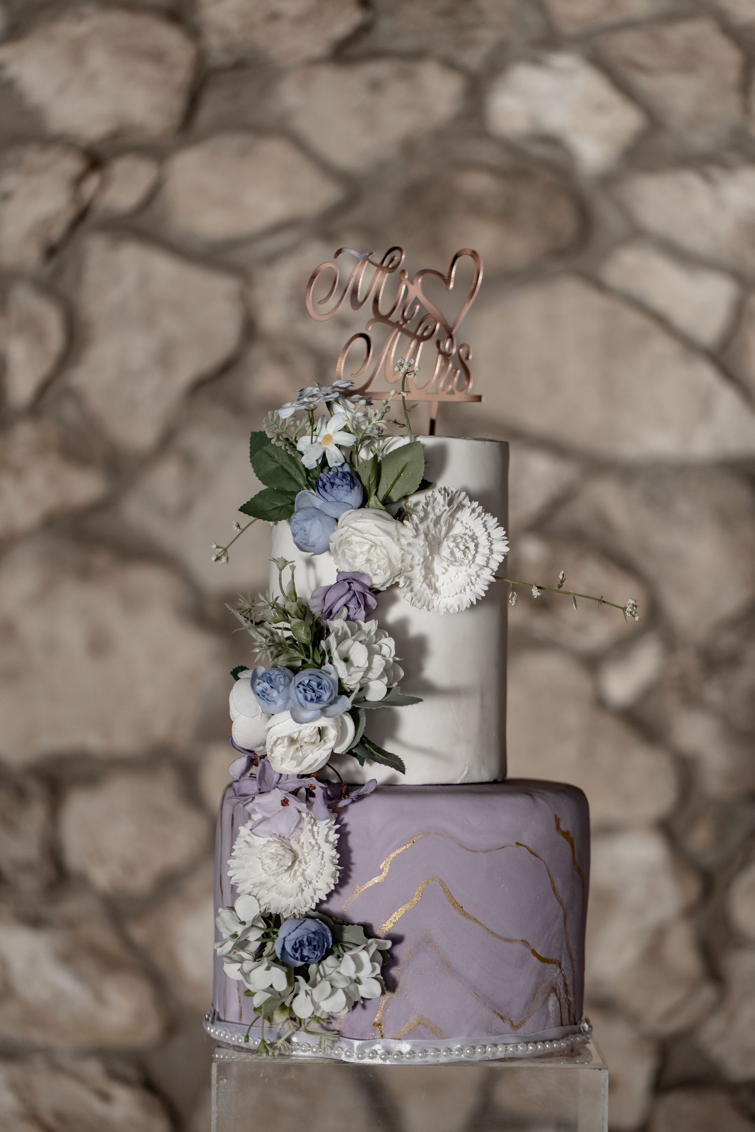 Elegant two-tiered wedding cake decorated with purple marble effect, metallic details and cascading blue and white flowers.