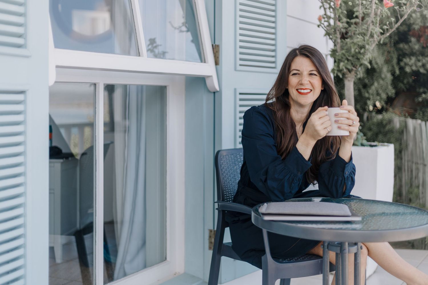 Person in navy blue clothing gestures by sunny windows with white shutters.