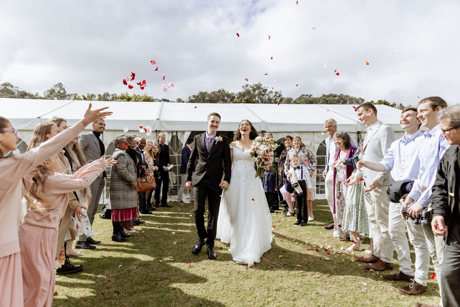 Wedding guests form a long receiving line on grassy lawn under bright sky for outdoor celebration.
