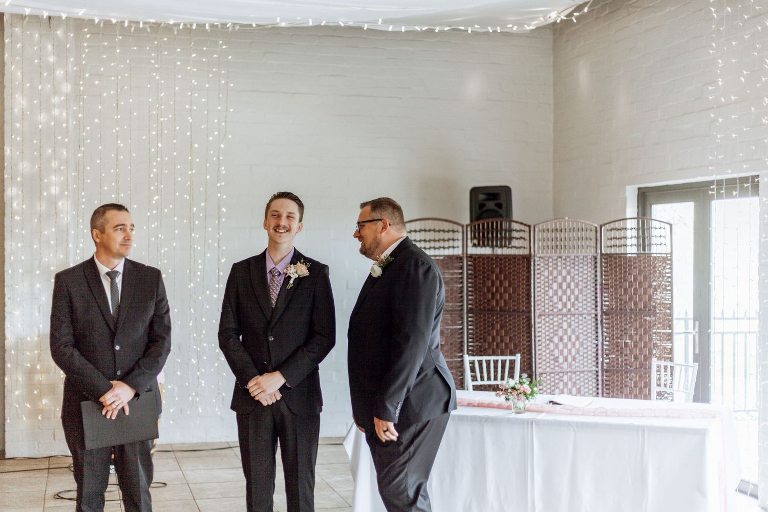 Three men in dark suits stand together in formal conversation against a white backdrop and latticed screens.