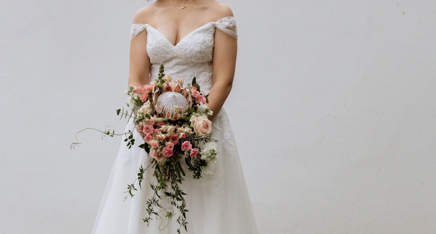 A bride in a strapless white wedding dress carries a cascading bouquet of pink flowers and greenery.