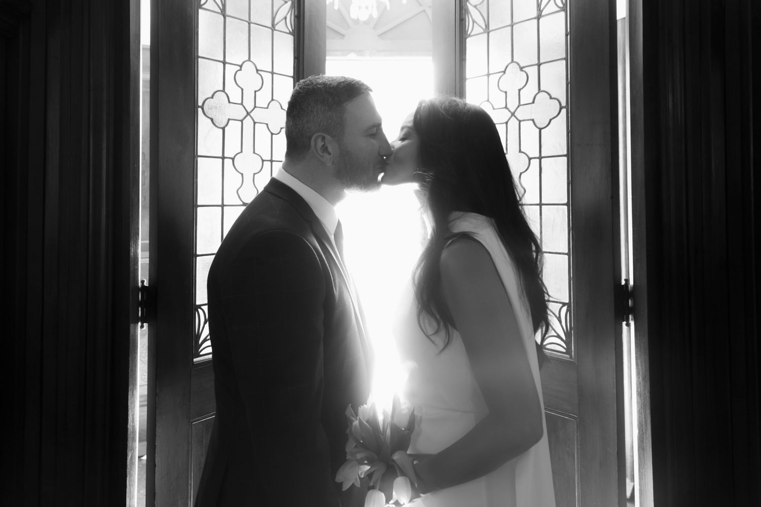 A romantic silhouette of a couple sharing a kiss in front of a decorative window with bright backlight.
