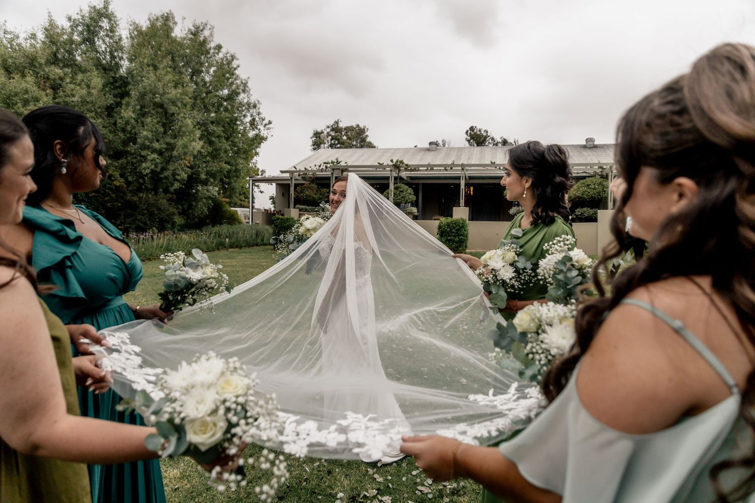 Bridesmaids in green dresses hold bride's floral veil at outdoor wedding venue.