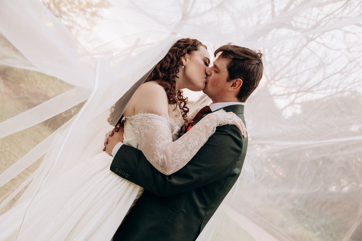 Bride and groom sharing a romantic kiss under a flowing veil on their wedding day.