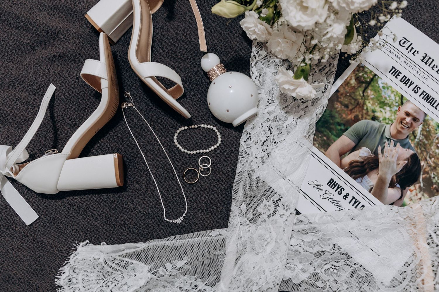Elegant wedding flat lay featuring white heels, pearl jewelry, rings, perfume bottle, lace veil, and a newspaper announcement.