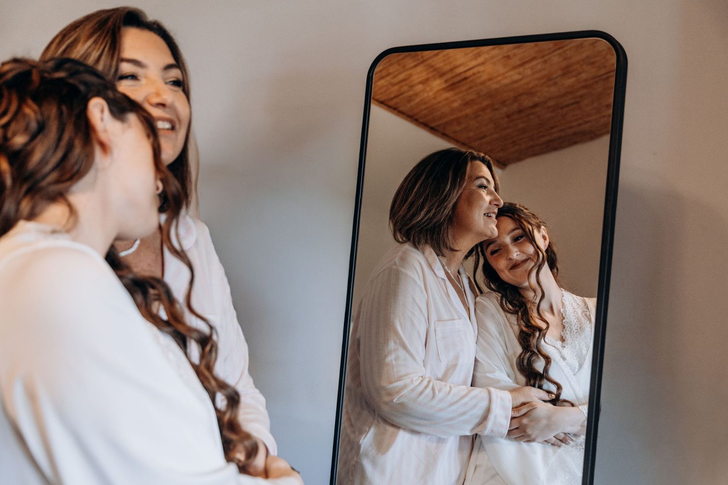 Two bridesmaids in white robes smile and embrace while viewing their reflection in a tall black-framed mirror.