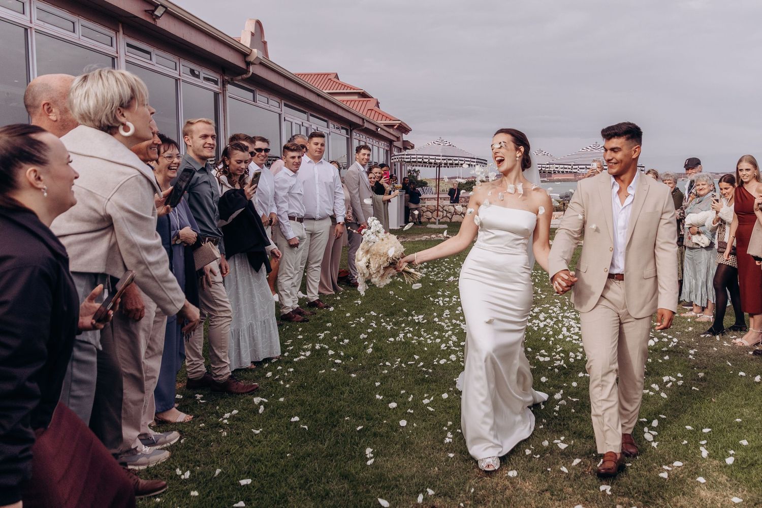Bride and groom walk through cheering guests outdoors at wedding reception venue near waterfront.