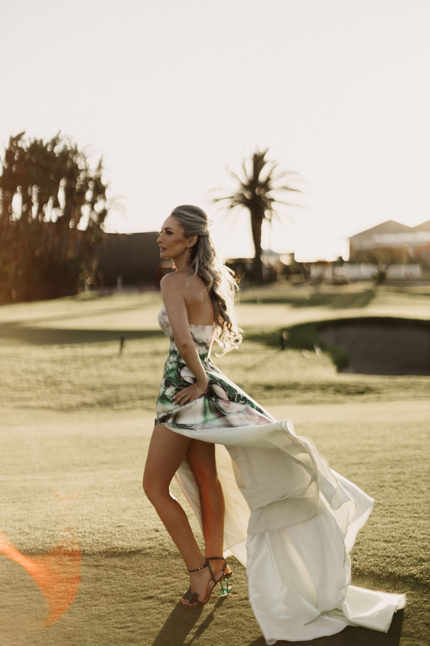 A bride in a flowing white dress twirls playfully on a sunlit golf course during golden hour.