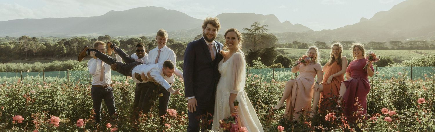 Panoramic view of a vineyard wedding ceremony setup with rustic wooden chairs and mountain backdrop at sunset.