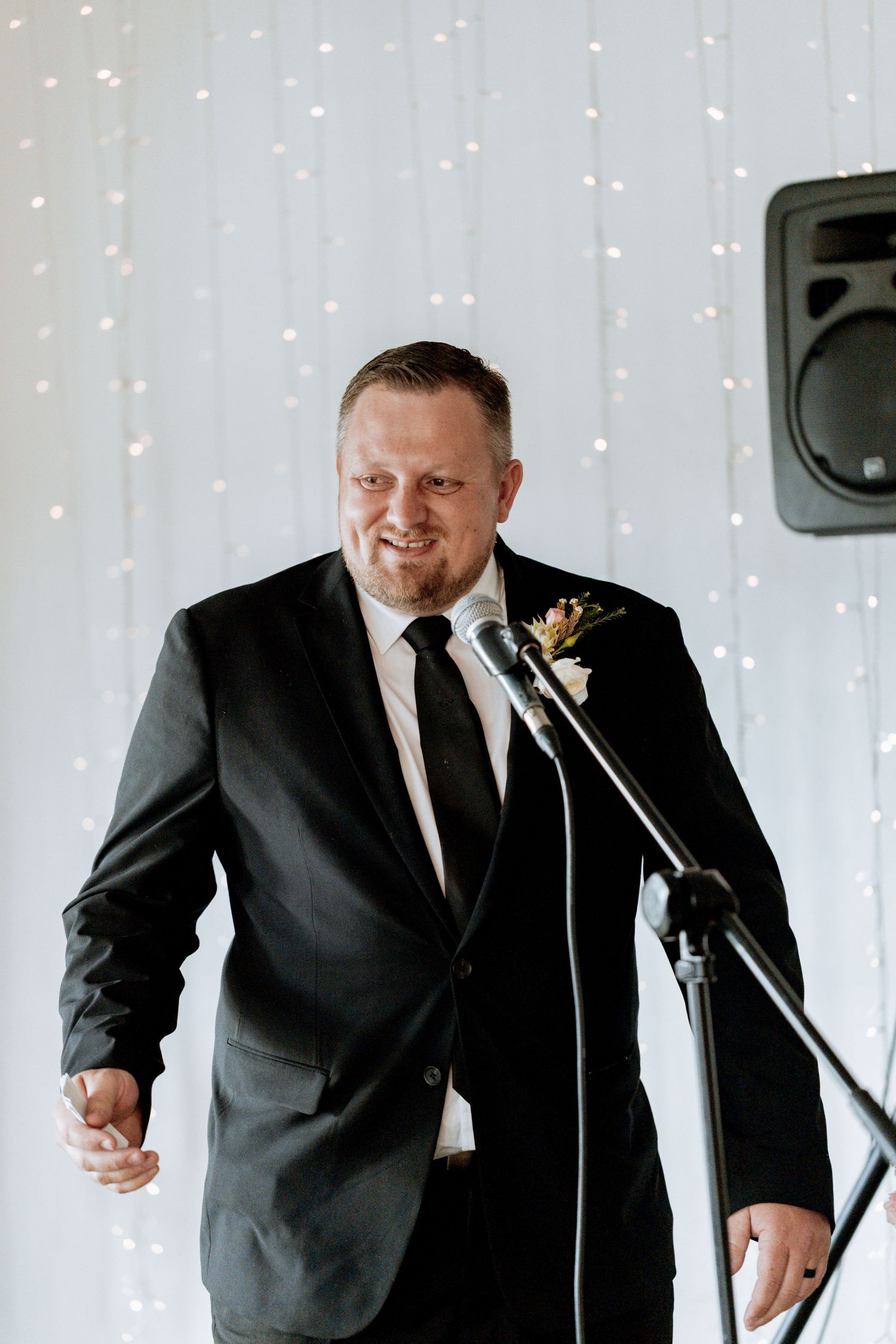 A person in a black suit and tie giving an animated speech at a podium with twinkling lights in the background.