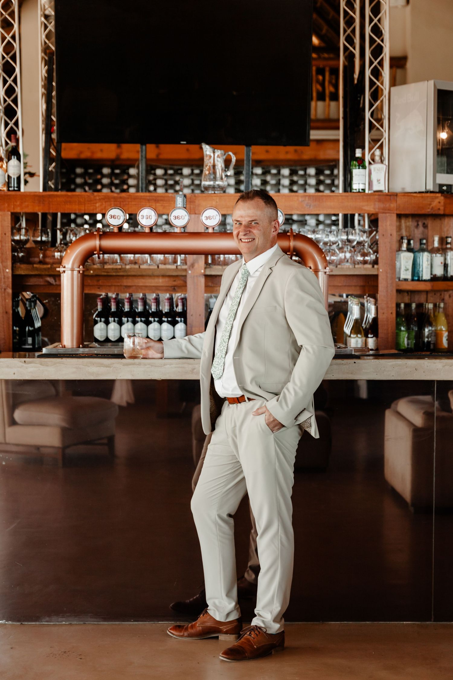 A person wearing a beige suit appears in multiple poses in front of a bar counter with wine bottles and glasses.