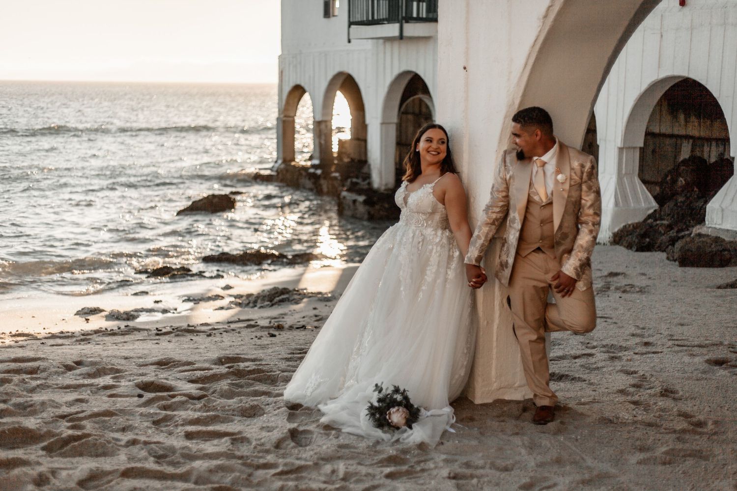 Couple in wedding attire walking along arched beachside colonnade at sunset with waves crashing on shore.