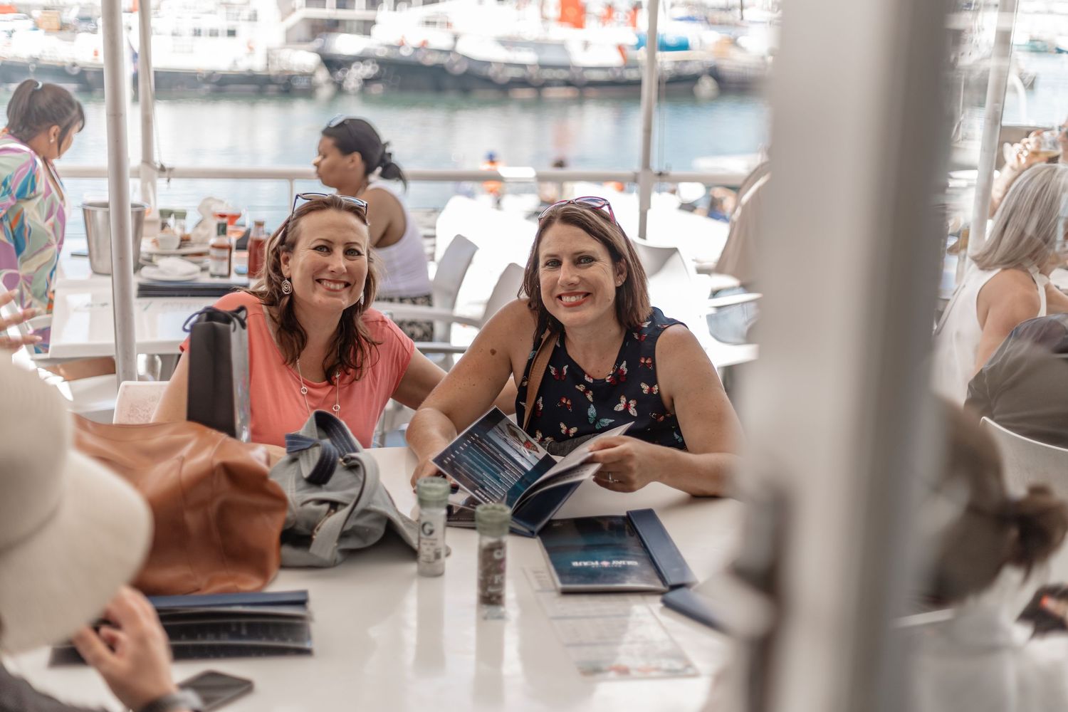 A group shares laughs and conversation around a table at an outdoor waterfront restaurant on a sunny day.