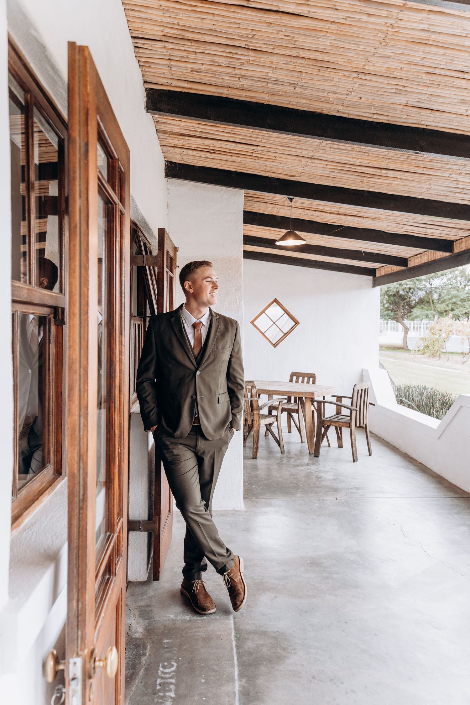 Smiling groom in gray suit leaning against wall on covered patio with wooden ceiling and dining area.