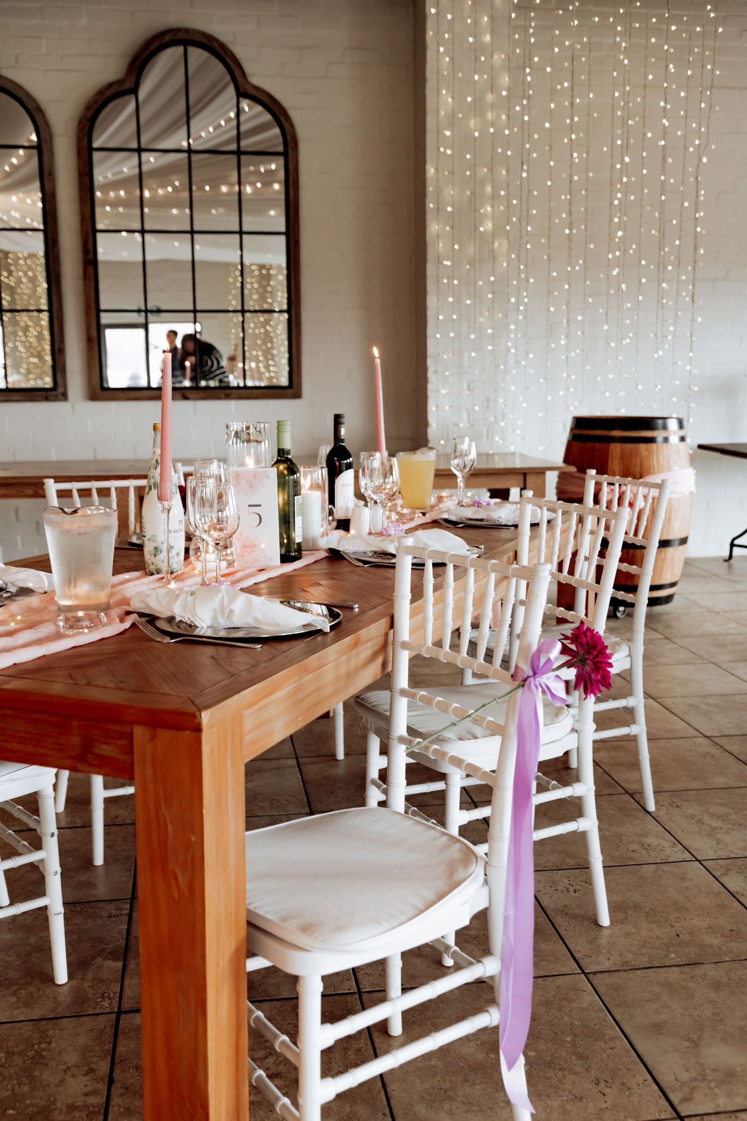 Long wooden table with white chairs decorated with pink ribbons and string lights create an elegant wedding reception setup.
