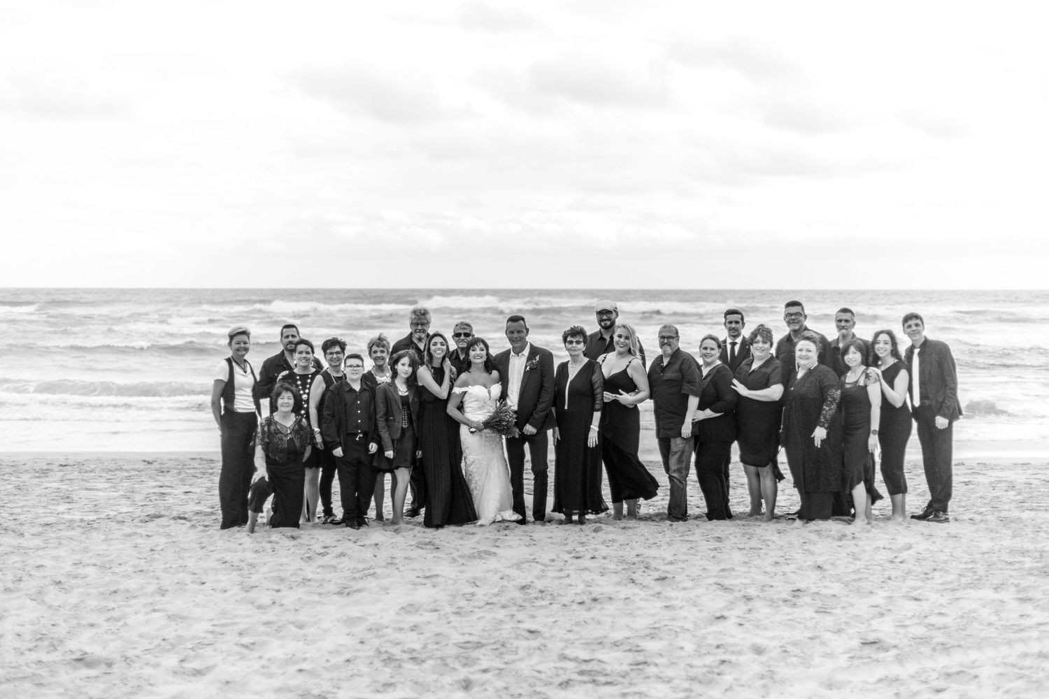 A large group of formally dressed people pose together on a beach with waves in the background.