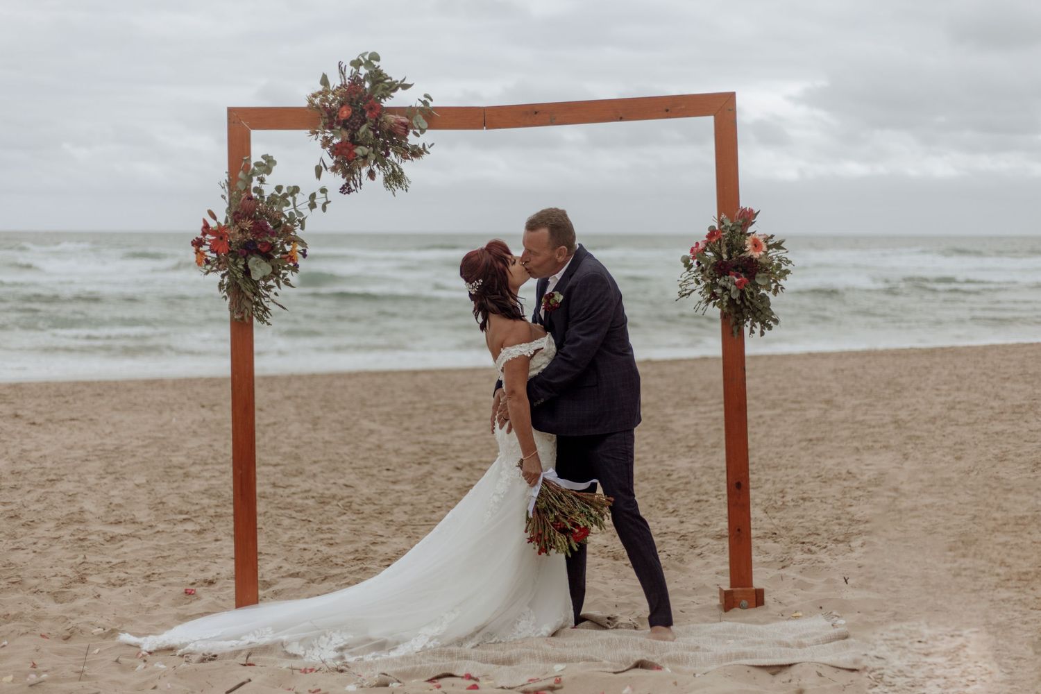 Rustic wooden frame decorated with flowers stands on a beach as waves crash in the background during a cloudy wedding day.