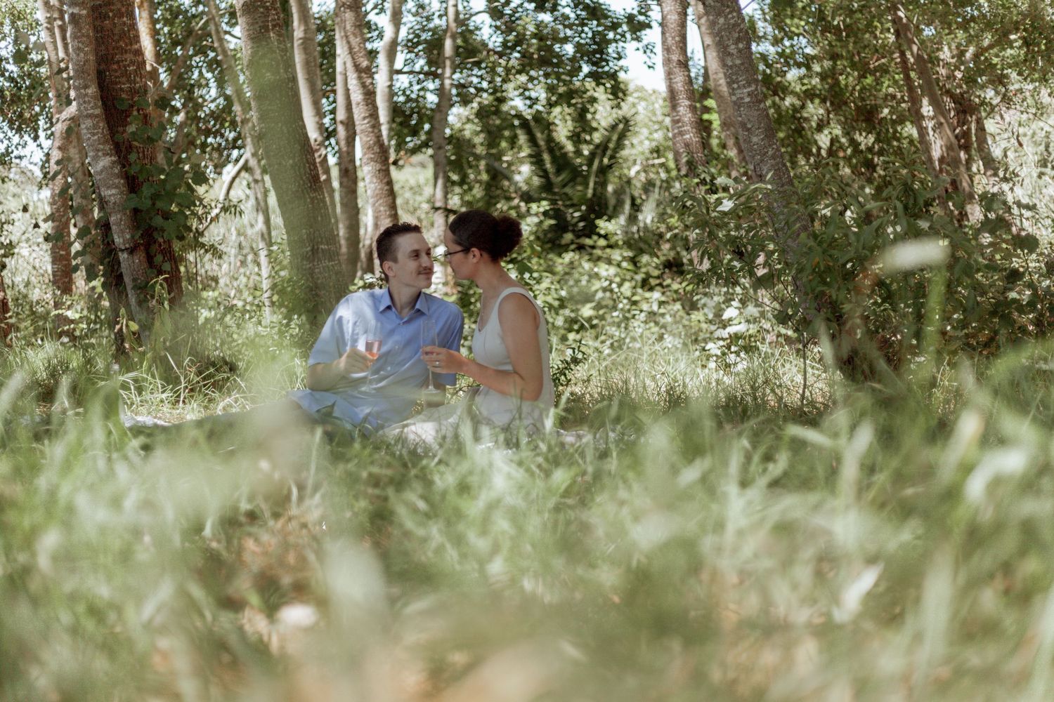 A couple enjoys a romantic picnic in a sun-dappled forest setting with wine and snacks.