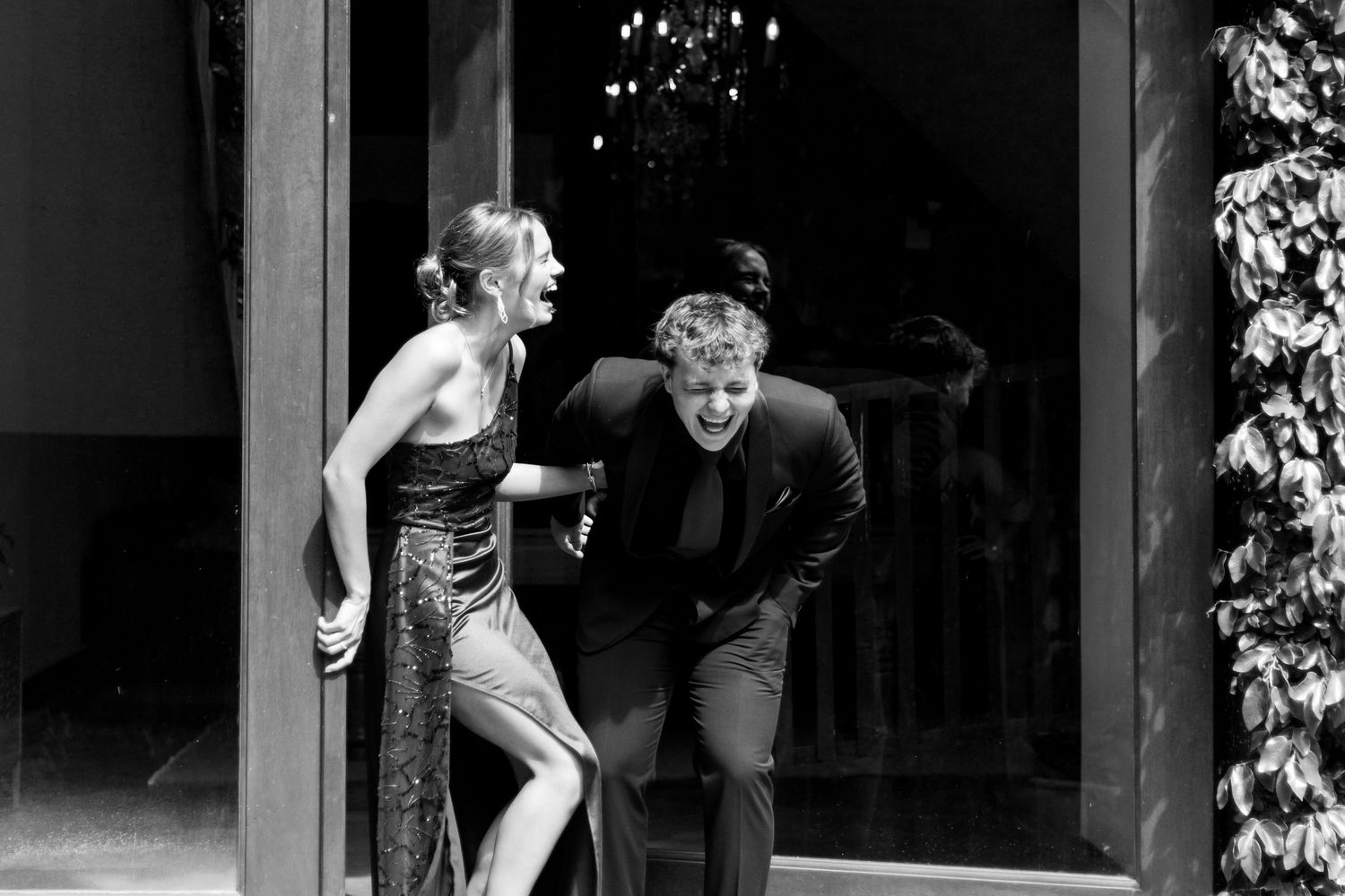 Black and white photo of two people laughing and leaning forward at a formal event near glass doors.