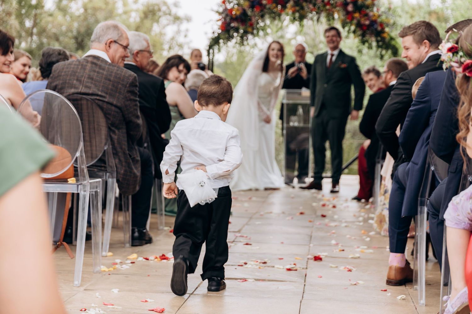 Young ring bearer walks down wedding aisle scattering rose petals as bride and groom stand at floral altar.