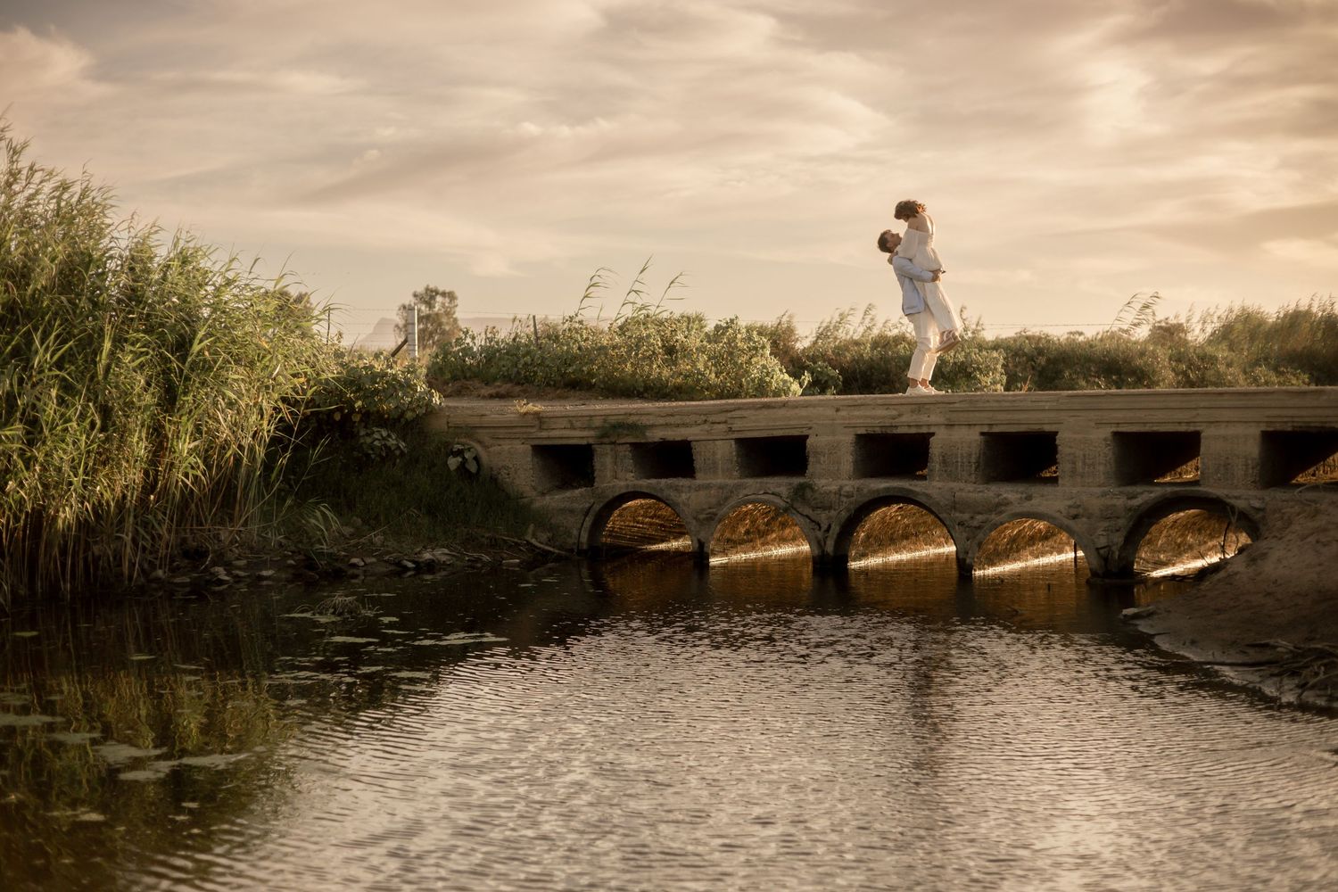 Couple embracing on a stone arch bridge over calm water surrounded by tall reeds under a dramatic cloudy sky.